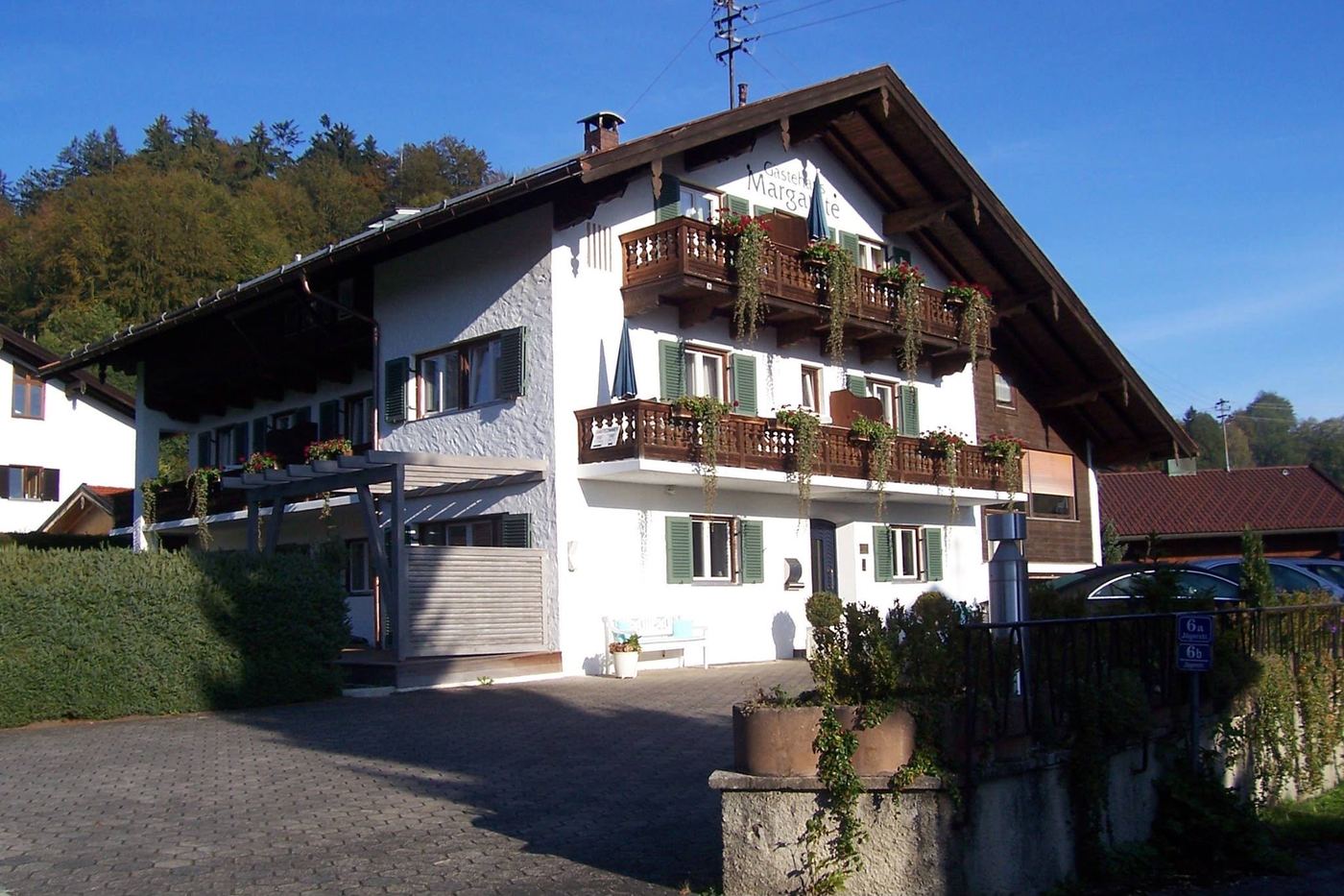 Das Gästehaus Margarete von der Straße aus: weißes Haus mit
              Holzbalkonen, roten Geranien und dem Schriftzug am Giebel